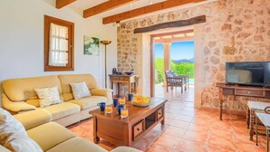 A cozy living room with a sofa, coffee table, and TV. There's a stone wall and a view of greenery outside. - Villa Can Xanet de Baix by Villa Plus (Mallorca)