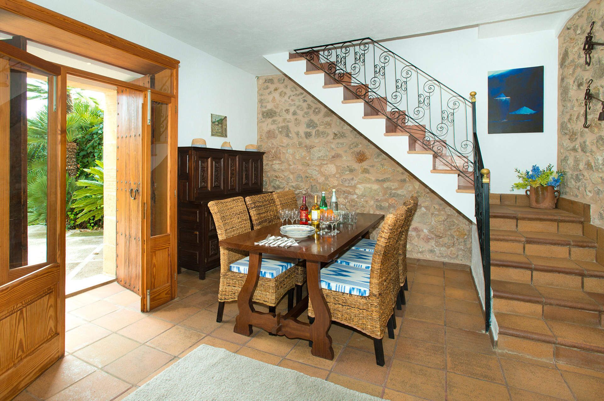 A dining area with a wooden table and wicker chairs, a stone wall, and a staircase with a wrought iron railing.