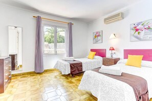 A room with two beds, a window with curtains, a bedside table with a lamp, and a wall-mounted air conditioner.