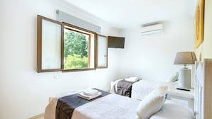 Ein Hotelzimmer mit großem Fenster, einem Bett mit weißer Bettdecke, einem Nachttisch mit Lampe und einem an der Wand montierten Fernseher.