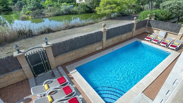 Ein rechteckiger Pool mit klarem, blauem Wasser, umgeben von einer Steinmauer und einer Holzterrasse. Liegestühle mit roten Kissen und eine kleine Gartenanlage mit Teich und Bäumen sind ebenfalls vorhanden.