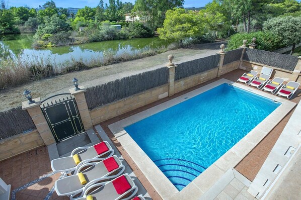 A rectangular pool with clear blue water, surrounded by a stone wall and a wooden deck. There are lounge chairs with red cushions and a small garden area with a pond and trees.