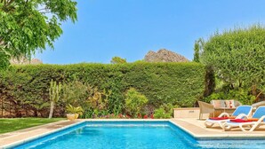 Ein rechteckiger Pool mit klarem, blauem Wasser, umgeben von einem gepflegten Garten mit verschiedenen Pflanzen und einer Hecke. Liegestühle und ein Rattansofa mit Kissen sind vorhanden.