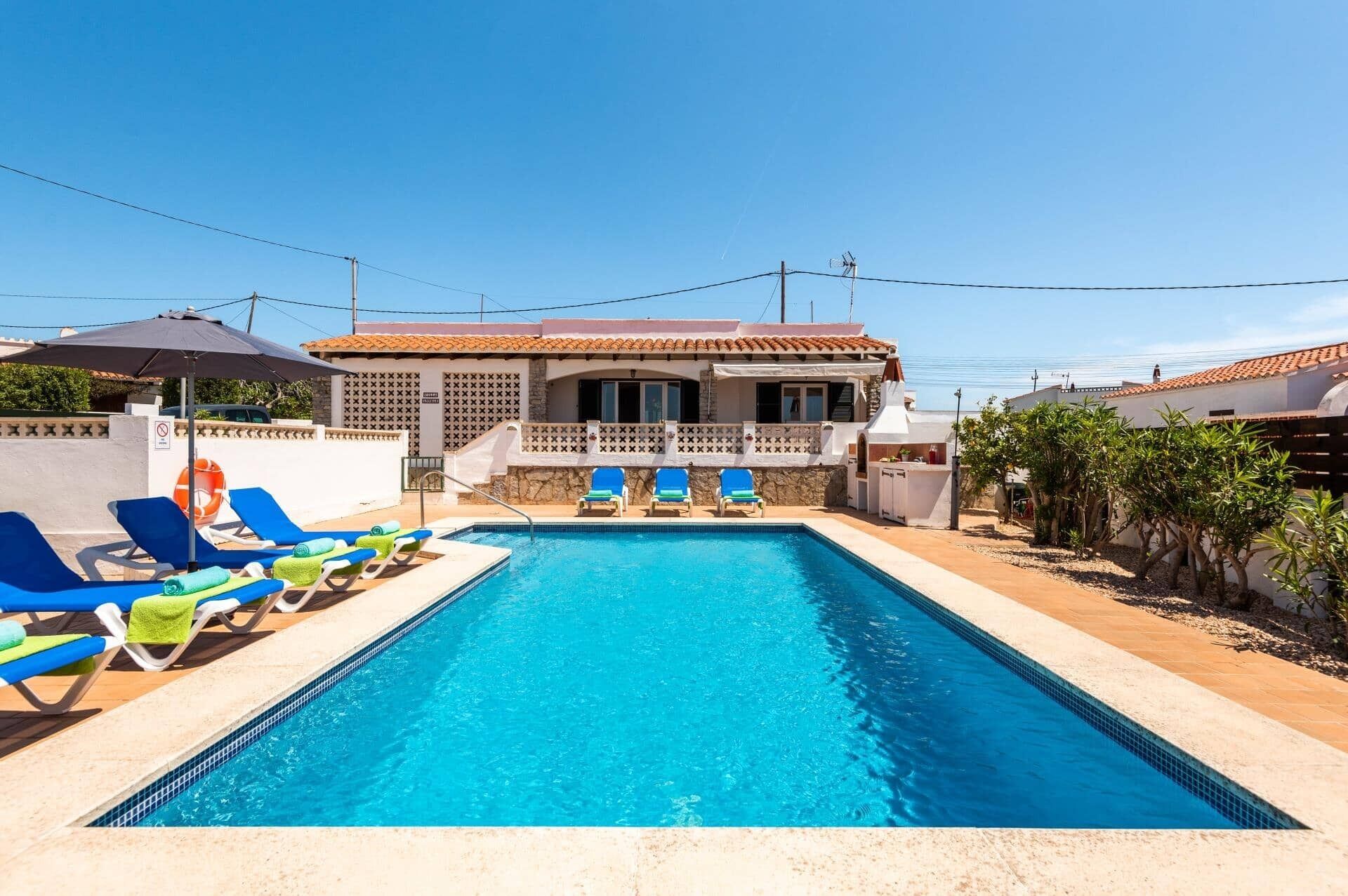 A swimming pool with blue lounge chairs and a building with a tiled roof in the background.