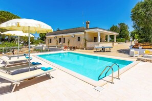 Una piscina con lettini e ombrelloni, un'area relax coperta e un edificio in pietra con un camino.
