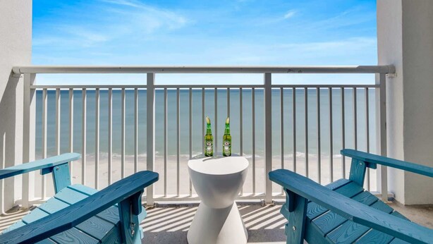 Ein Balkon mit weißer Reling, zwei blauen Stühlen und einem weißen Tisch mit zwei Flaschen darauf.