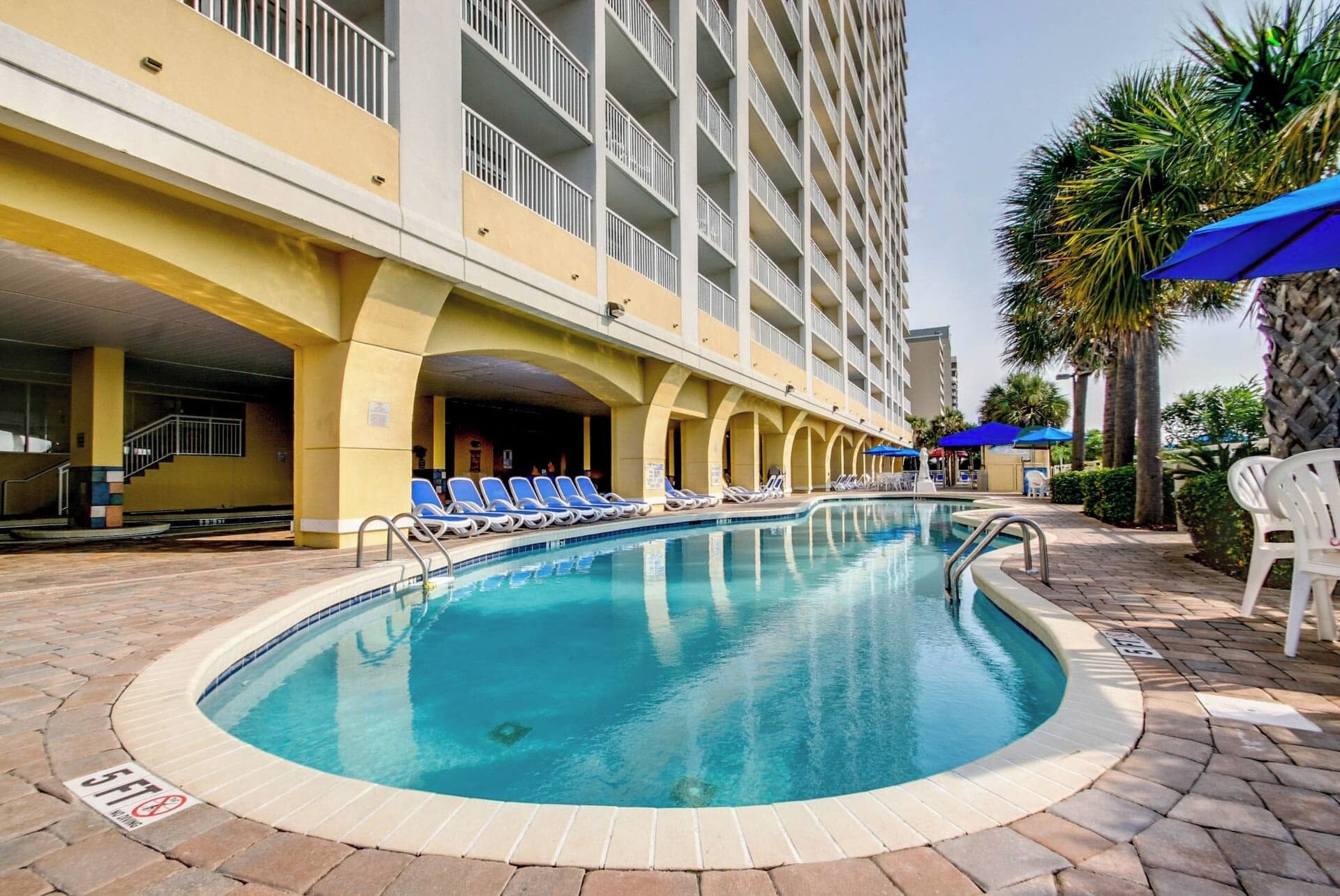 A swimming pool with lounge chairs, a building with balconies, and palm trees.