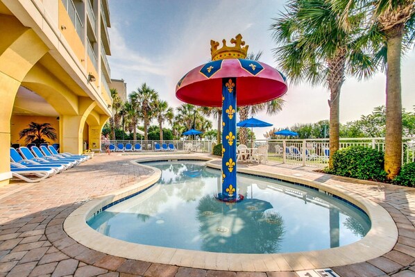 A pool area with a central fountain, lounge chairs, and palm trees. - Ocean Breeze by StayMareni - King Suite (Myrtle Beach)
