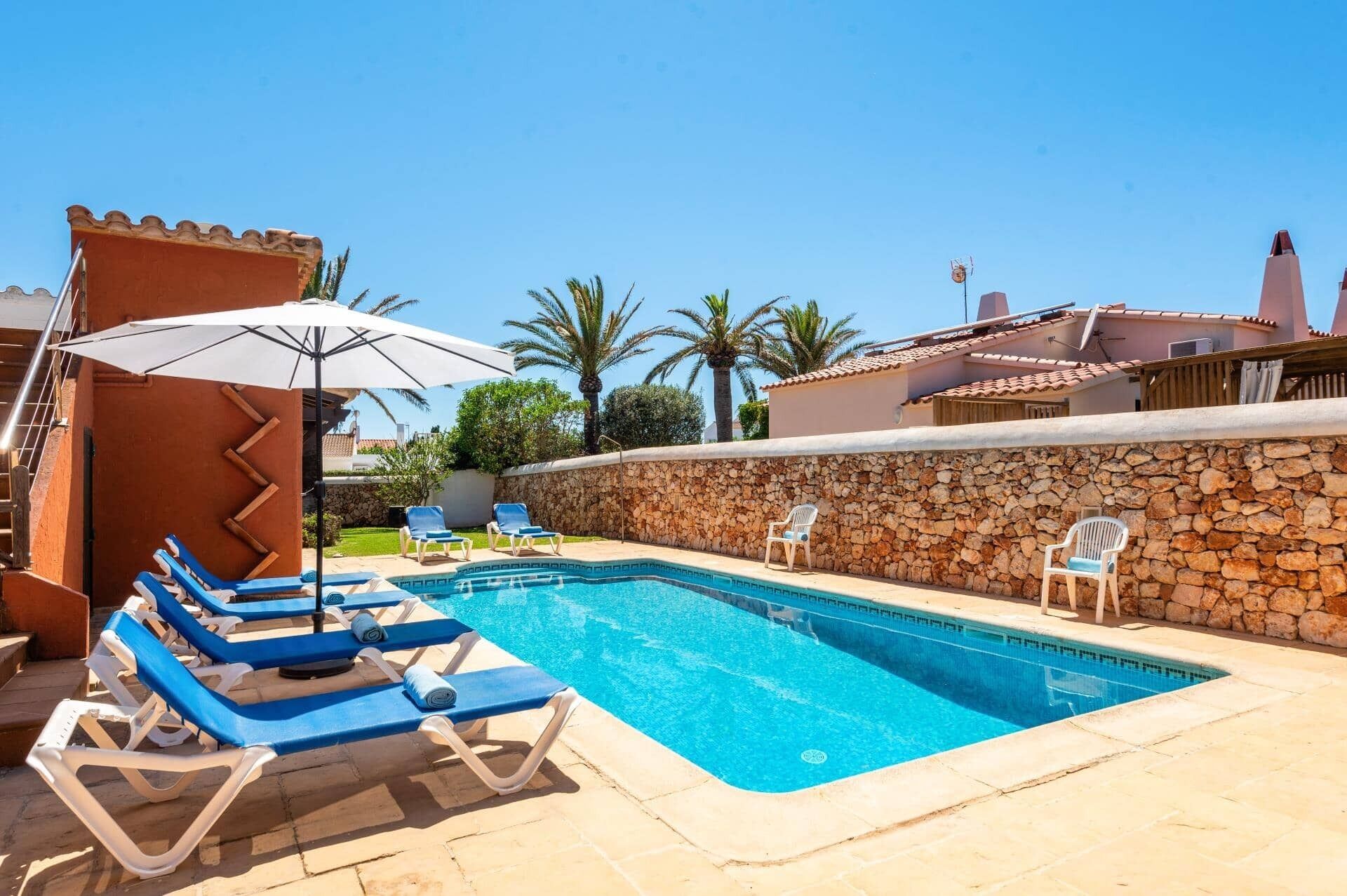 Une piscine entourée d’un mur de pierre et de palmiers, avec des chaises longues et des parasols.