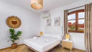 Een slaapkamer met een bed, nachtkastjes, een kamerplant, een raam met gordijnen en een wandairconditioning.