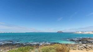 A coastal landscape with clear blue water, rocky shoreline, and distant buildings. - Villa Atlantico by Villa Plus (Corralejo)