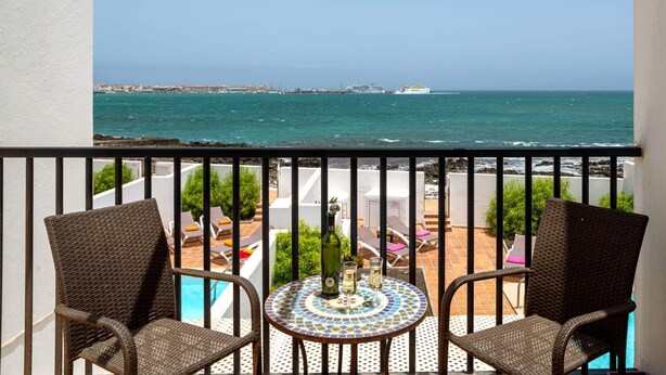 A balcony with wicker chairs, a tiled table, and a view of the sea and distant buildings. - Villa Bella Vista by Villa Plus (Corralejo)