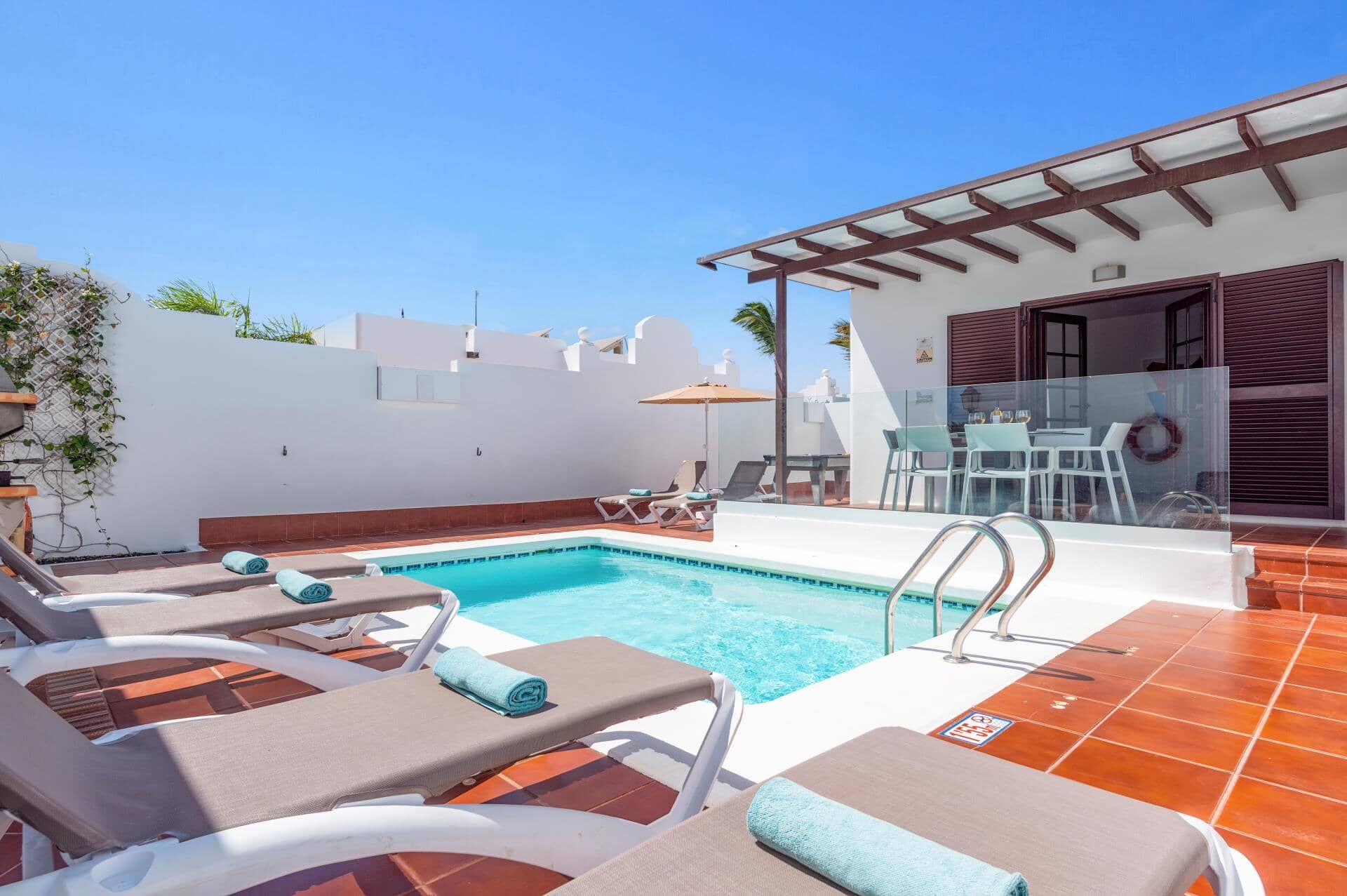 Un espace piscine aménagé avec des chaises longues et une table, entouré de murs blancs et protégé par une paroi en verre.