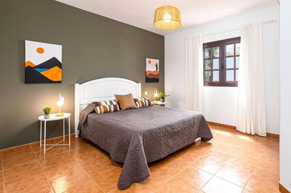 A bedroom with a bed, a bedside table with a lamp, a wall art piece, a window with curtains, and a hanging light fixture. - Villa Denise by Villa Plus (Tías)