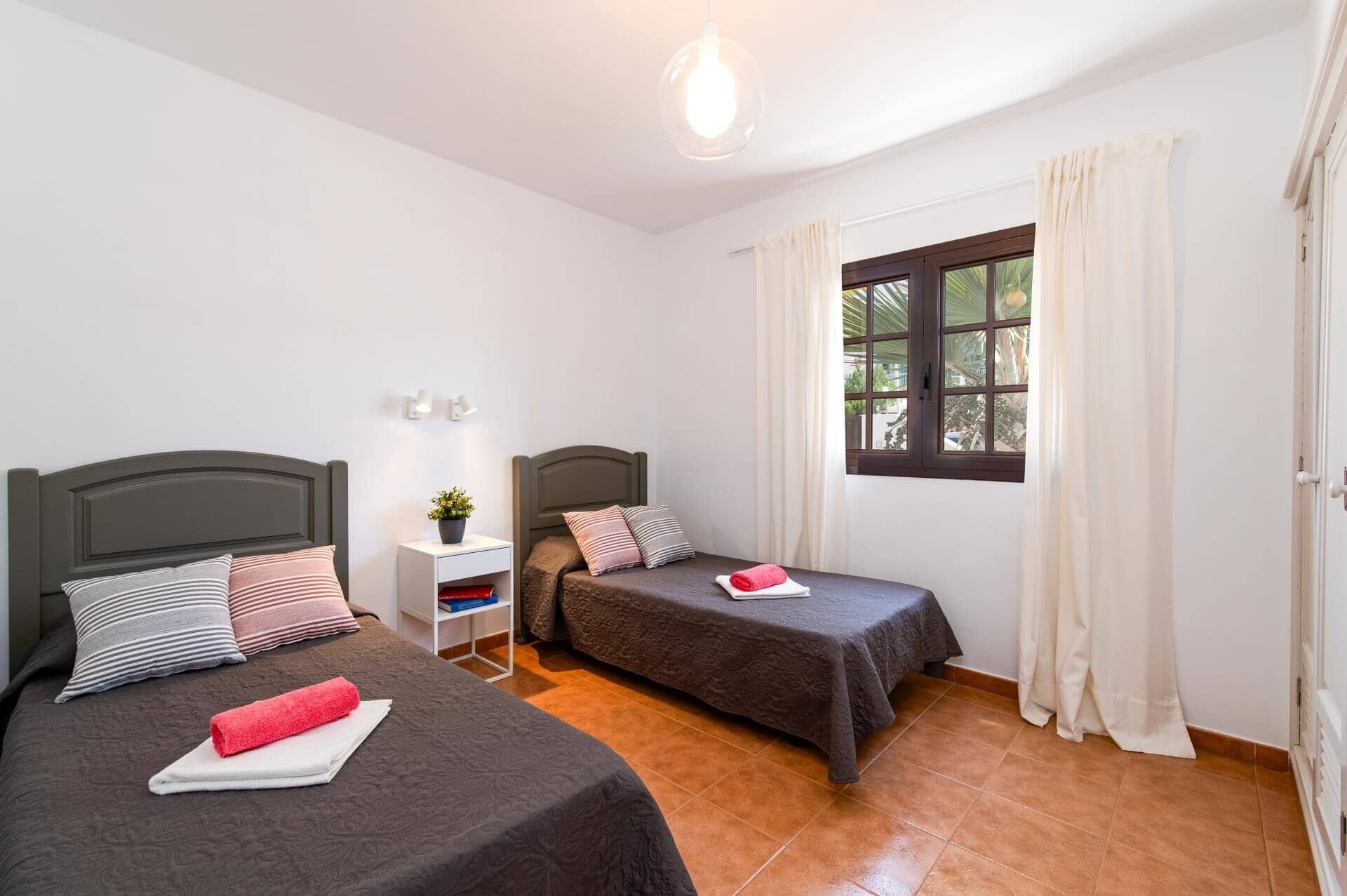 A room with two beds, a small bedside table with a plant, a window with curtains, and a light fixture.