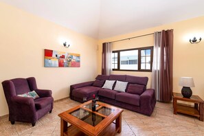 Una sala de estar con un sofá modular, un sillón individual, una mesa de centro de vidrio, una lámpara y un cuadro en la pared.