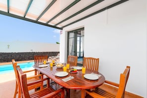 A wooden dining table with chairs, plates, cutlery, and glasses of orange juice, set against a poolside backdrop.