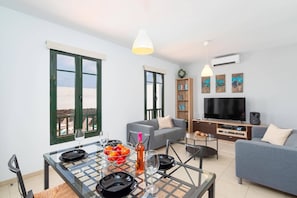 A modern living room with a dining area, a grey sofa, a flat-screen TV, and a bookshelf.