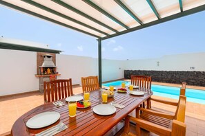 An outdoor dining area with a pool, wooden table, chairs, and a grill. - Villa Celeste Lanzarote by Villa Plus (Playa Blanca)