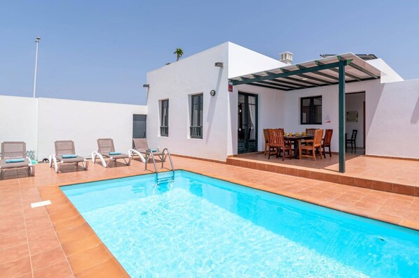 A swimming pool with lounge chairs, a covered outdoor dining area, and a white building with windows. - Villa Celeste Lanzarote by Villa Plus (Playa Blanca)