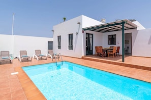 A swimming pool with lounge chairs, a covered outdoor dining area, and a white building with windows.