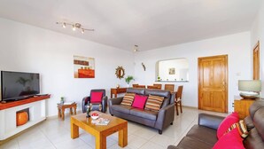 A modern living room with a flat-screen TV, a wooden coffee table, a grey sofa with striped cushions, and a dining area with a wooden table and chairs. - Villa Casa Linden by Villa Plus (Playa Blanca)