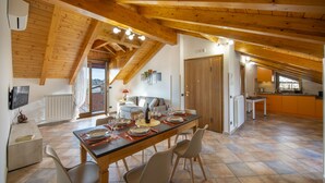 A spacious dining area with a long table set for six, wooden beams on the ceiling, a kitchen in the background, and a view of the outdoors. - Penthouse with Terrace—Hakuna Matata in Sentieri (Agerola)