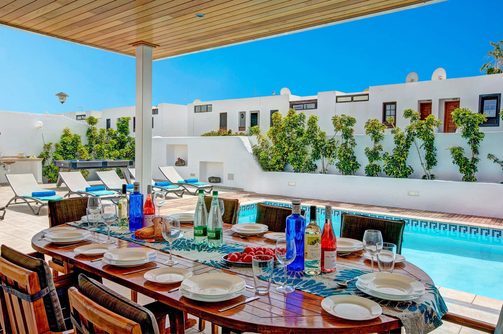 An outdoor dining area with a pool view, white buildings, and greenery.