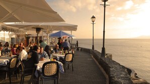 A seaside restaurant with outdoor seating, a view of the sea, and a stone wall along the walkway.
