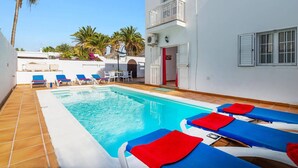 A swimming pool with blue and red lounge chairs, a white building with a balcony, and palm trees in the background.