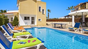 A swimming pool with lounge chairs and a table, surrounded by buildings and palm trees. - Villa Azalea by Villa Plus (Peyia)