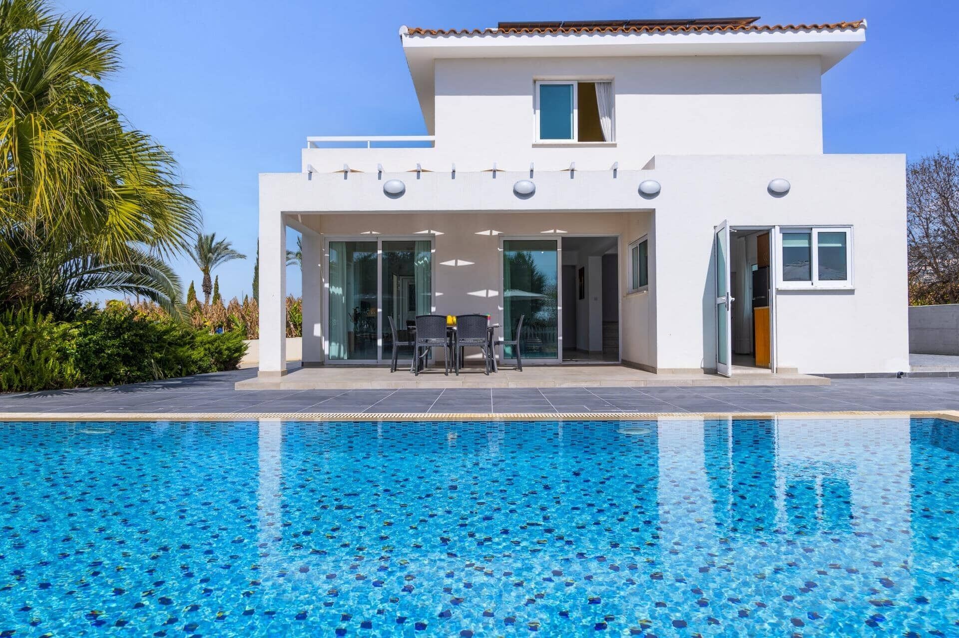 Ein modernes Haus mit Swimmingpool, einer überdachten Terrasse und Gartenmöbeln, und einem strahlend blauen Himmel.