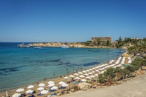 A beach with white umbrellas and lounge chairs, a building with multiple floors, and boats in the water.