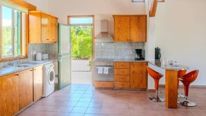 A kitchen with wooden cabinets, a washing machine, a coffee maker, and two bar stools. - Villa Ibiscus by Villa Plus (Poli Crysochous)