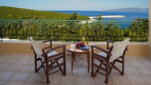 A balcony with two wooden chairs, a small round table, and a view of the sea and greenery.