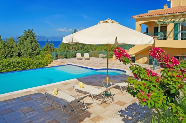 A swimming pool with lounge chairs and an umbrella, surrounded by greenery and a view of the sea.