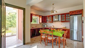 A kitchen with a dining table, chairs, a refrigerator, and a window overlooking greenery. - Villa Chrissos by Villa Plus (Lourdata)