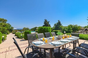 An outdoor dining area with a table set for four, surrounded by chairs, with a view of the sea and a barbecue grill in the background.