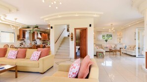 A spacious living area with a sofa set, a coffee table, and a dining table. A kitchen is visible in the background, and there is a staircase leading to an upper level.