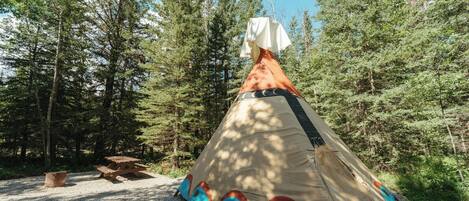 A conical tent with a white covering, set up on a gravel surface surrounded by trees.
