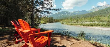 A river with a forested mountain backdrop, red Adirondack chairs, and a clear sky.