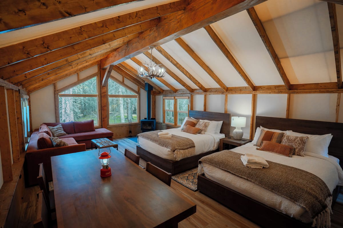 A cozy attic bedroom with a wooden ceiling, two beds, a sofa, and a dining table.