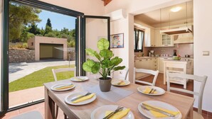 A dining area with a table set for four, a potted plant, and a view of the garden and garage through glass doors. - Villa Dimitra Springs by Villa Plus (Rhodes)