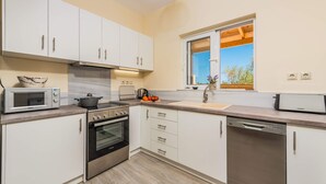 A modern kitchen with white cabinets, a built-in oven, a microwave, a dishwasher, and a window with a view of trees.