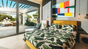 Une chambre moderne avec un grand lit, une vue sur l’extérieur, un tableau abstrait aux couleurs vives et une petite table avec une chaise.