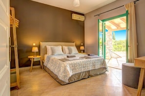 A bedroom with a bed, two bedside tables, a nightstand, a window with curtains, and a view of greenery outside.