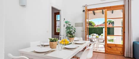 A dining area with a white table set for four, surrounded by white chairs, with a view of a garden through a glass door.