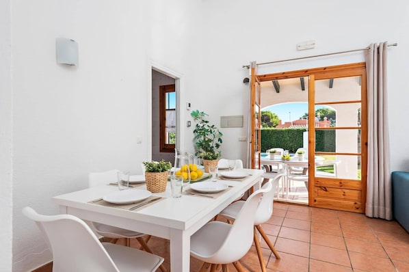 A dining area with a white table set for four, surrounded by white chairs, with a view of a garden through a glass door. - Villa Belen by Villa Plus (Cap d'Artrutx)