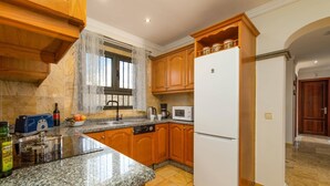 Uma cozinha com bancadas de granito, armários de madeira, uma geladeira branca e uma janela com cortinas de renda.