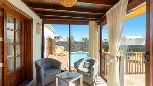 Uma área externa coberta com dois sofás cinzentos, uma mesa pequena com um livro e um copo de vinho, e vista para uma piscina e um muro de pedra.